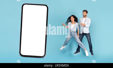 Shocked glad young caucasian man and arab lady jump in air, point finger at phone with blank space Stock Photo