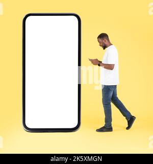 African Guy Using Cellphone Walking Near Large Phone, Yellow Background Stock Photo