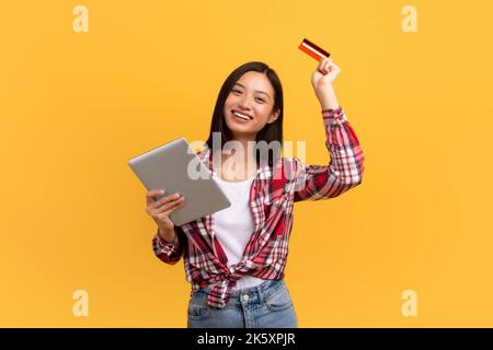 Online shopping concept. Happy japanese lady using digital tablet and credit card, standing over yellow background Stock Photo