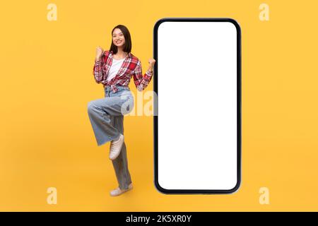 Great mobile offer. Overjoyed asian lady jumping near big smartphone with empty screen, celebrating success, mockup Stock Photo