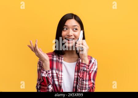 Cheerful positive asian lady in casual gesturing and calling on smartphone, isolated on yellow background, studio shot Stock Photo