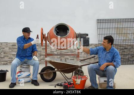 Two construction workers one young and one elderly during their lunch break while toasting with a beer. Smiling workers on a construction site surroun Stock Photo