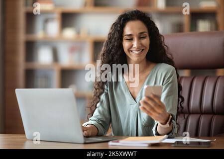 Happy middle eastern woman sitting at workdesk, using smartphone Stock Photo