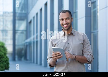 Portrait of a happy businessman outside an office building, a man in a casual shirt is looking at the camera and smiling, a businessman is using a tablet computer. Stock Photo