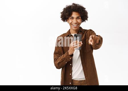 Portrait of gen-z guy pointing at camera, recording video or taking photo on smartphone camera, smiling and laughing, standing over white background Stock Photo