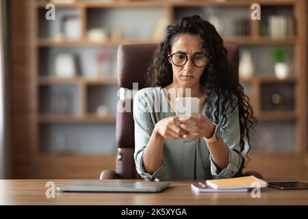 Concentrated woman entrepreneur sitting at workplace, using smartphone Stock Photo