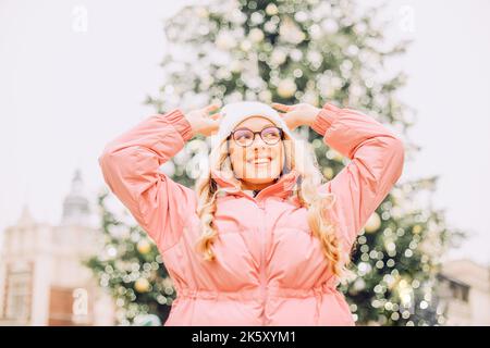 Cute portrait of a girl in glasses on the background of a Christmas tree. Winter, the first snow, a blonde in a pink jacket and a white hat smiles. Ne Stock Photo