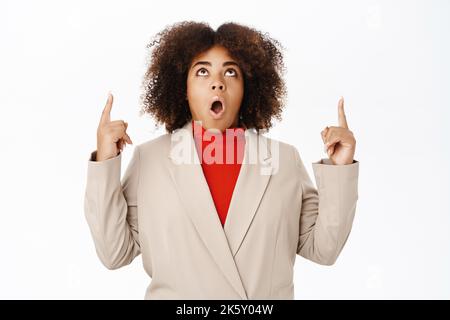 Portrait of surprised black woman, saleswoman pointing fingers up, looks impressed and amazed by company banner, standing over white background Stock Photo