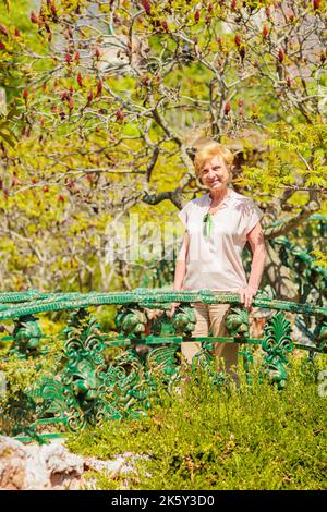 Senior woman standing on a beautiful forged bridge surrounded by greenery Stock Photo