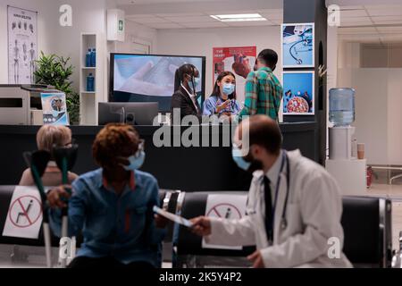 Female hospital staff members attending a sick young african american man at reception area. Doctor handing prescription to woman with partial body paralysis. Medical clinic lobby during covid pandemic. Stock Photo