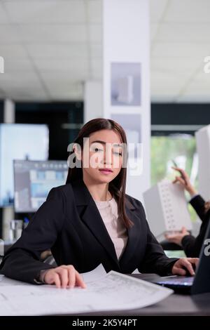 Asian architect working with project blueprints in coworking space, asian employee in architecture office at workplace desk. Real estate agency worker, software developer reading plan Stock Photo