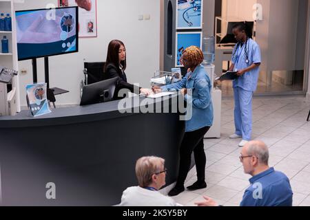 Young adult filling in registration form at hospital reception desk, writing medical checkup report before attending appointment. Woman checking papers in facility lobby, having consultation. Stock Photo