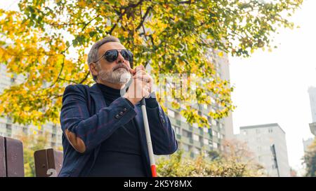 Outdoor medium closeup shot of 60-year-old bearded caucasian man with complete loss of sight dressed in blazer, sitting on bench at park, holding walking cane with two hands and relaxing in fresh air. High quality photo Stock Photo