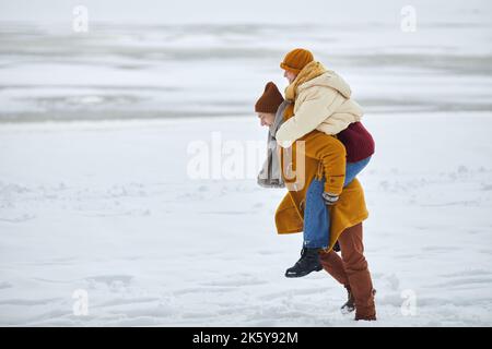 Side view portrait of young man carrying girlfriend on shoulders while having fun outdoors in minimal winter landscape, copy space Stock Photo