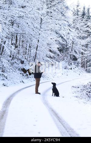 Black dog sitting on ground looks at man holding stick on a snow covered road in the Bavarian Alps on a cold autumn day in Germany. Stock Photo