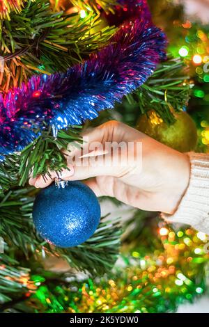 Female hands hanging Christmas ball on Christmas tree. Woman decorates artificial festive tree with colored tinsel and garland. Concept of eco-friendl Stock Photo