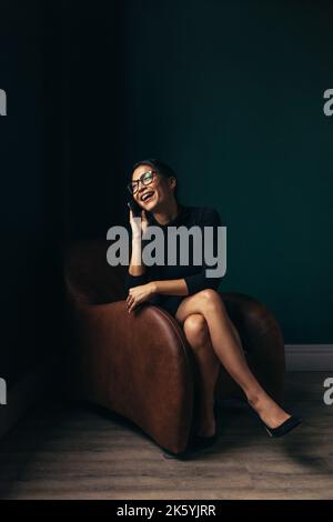 Young woman talking on the phone and smiling happily. Beautiful asian woman taking a phone call while sitting in an armchair. Stock Photo