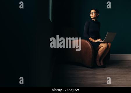 Thoughtful lady sitting on armchair with laptop and looking away. Young woman working on laptop and looking outside window. Stock Photo