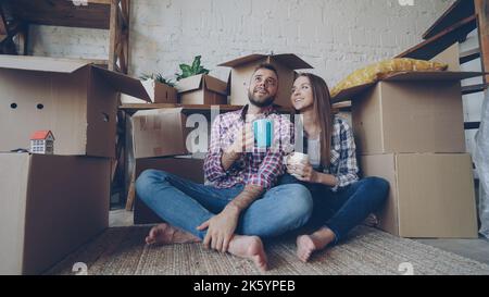 Young husband and wife are talking, looking around sitting on floor after moving to new house. Nice interior and many boxes with personal things are visible. Stock Photo