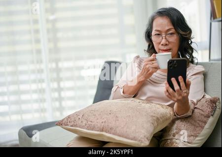 Concentrated 60s retired asian woman wearing glasses using her smartphone to read an online news while sipping coffee in the living room. Stock Photo