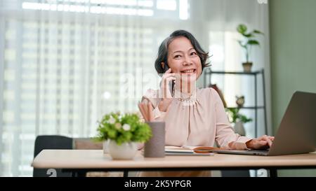Charming and cheerful aged-asian businesswoman or female executive manager enjoys talking on the phone while working in her office. Stock Photo