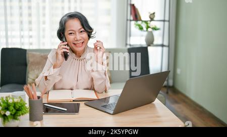 Charming and cheerful senior asian businesswoman talking on the phone with her business supplier while working at her desk in the office. Stock Photo