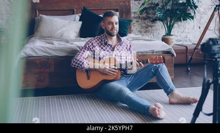 Handsome man blogger and guitarist is recording video lesson about guitar in bedroom using camera. Bearded guy is greeting his followers, talking and showing guitar parts. Stock Photo