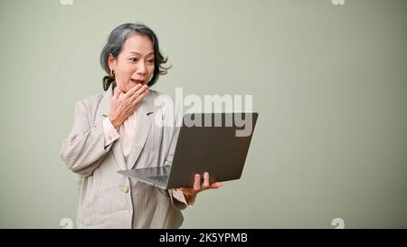 Amazed and surprised 60s aged-asian woman or businesswoman holding and looking at laptop screen, standing isolated over green background. shocked, exc Stock Photo