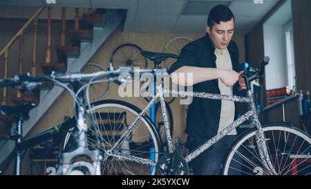Experienced male master is checking and repairing bicycle handle-bar with special tools while working in workshop with numerous spare parts and equipment. Stock Photo