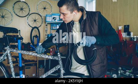 Young mechanic owner of bike repairing workshop is fixing broken bicycle holding bundle of wire and fixing it to bike frame. Small business and maintenance concept. Stock Photo