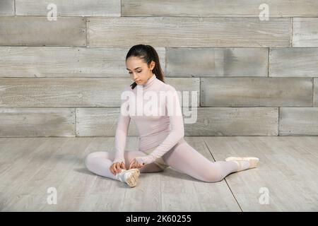Full body of young slim female in bodysuit sitting on floor and fixing ballet shoes before dance session Stock Photo