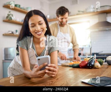 Happy young woman browsing social media on smartphone while her boyfriend cooks in the background. Young hispanic female searching for recipe on Stock Photo