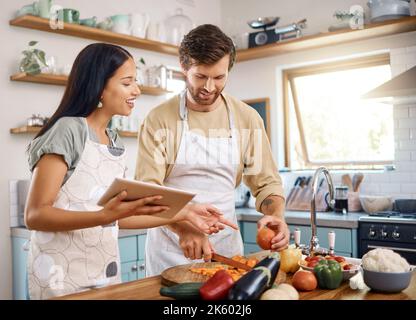 Happy young interracial couple using digital tablet while cooking together at home. Young man cutting vegetables while wife explains what to do while Stock Photo