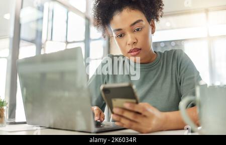 Young serious mixed race woman working a laptop and using a phone sitting at a table at home. One focused hispanic female with a curly afro planning Stock Photo