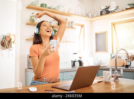 Young beautiful happy mixed race businesswoman holding and using a phone while wearing headphones and listening to music working from home. One joyful Stock Photo