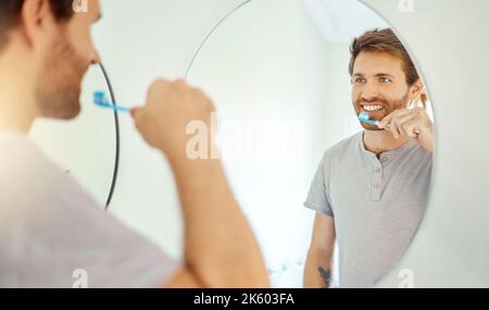 One handsome man brushing his teeth in a bathroom at home. Caucasian male using whitening toothpaste and looking in a mirror in his apartment. Stock Photo