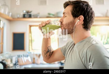 One fit young caucasian man drinking a glass of healthy green detox smoothie while wearing earphones in a kitchen at home. Guy having fresh fruit Stock Photo