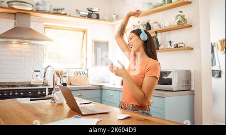 Young beautiful cheerful mixed race businesswoman holding and using a phone while wearing headphones and listening to music working from home. One Stock Photo