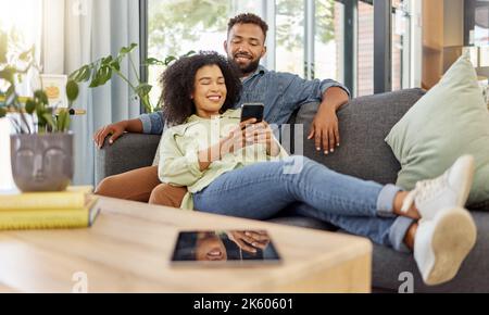 Happy young mixed race couple smiling while using a phone together at home. Cheerful hispanic boyfriend and girlfriend bonding and using social media Stock Photo