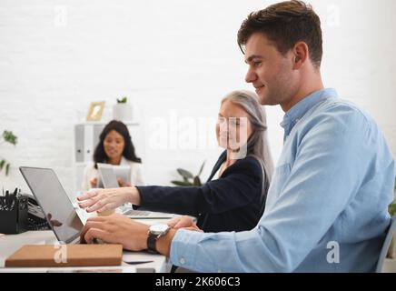 Mature caucasian businesswoman talking to a young male colleague while working on a laptop together at work. Boss showing an employee an idea on a Stock Photo