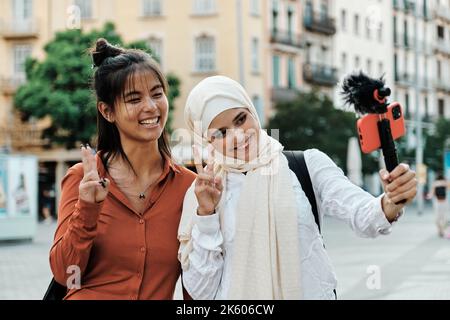 Multi-ethnic friends smiling and making peace sign with fingers while streaming with a mobile phone outdoors. Stock Photo