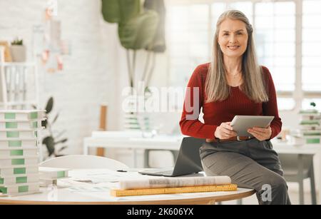 Mature businesswoman using a digital tablet. Senior boss sitting on her desk. Creative entrepreneur using an online wireless device in her agency Stock Photo
