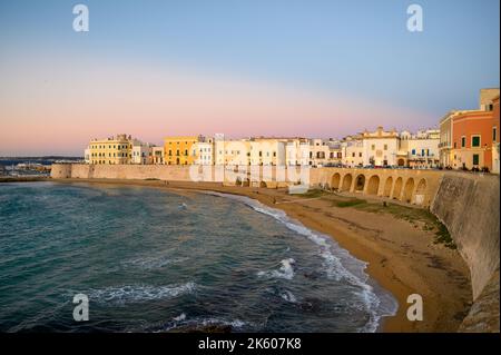 View across the bay and beach (Spiaggia della Purità) and waterfront facades of old town houses in evening sunlight in Gallipoli, Apulia, Italy. Stock Photo