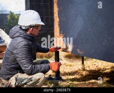 Male painter using paint roller, doing exterior paint work in a black color. Man worker building wooden frame house. Carpentry and construction concept. Stock Photo