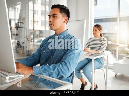 Young focused asian businessman using a desktop computer in an office at work. Serious chinese male businessperson typing an email at a desk. Business Stock Photo
