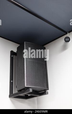 Close up of black big musical powerful speaker with protective grill hanging on wall in public place. Music speaker hangs on white wall near CCTV came Stock Photo