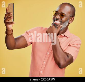 One happy trendy mature African American man taking selfies on his cellphone against a yellow studio background. Fashionable black man standing and Stock Photo