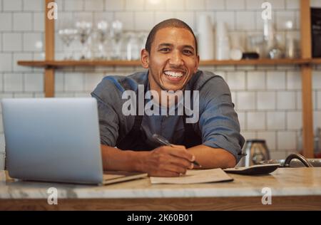 Portrait of business owner writing a list. Entrepreneur writing his goals in his cafe. Small business owner working on his laptop in his shop. Mixed Stock Photo