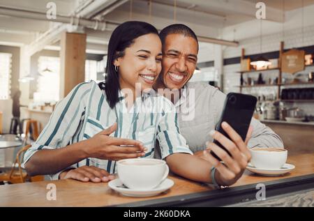Happy young mixed race couple sitting at table having coffee while looking at something on smartphone in cafe. Loving couple smiling while taking Stock Photo