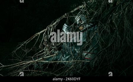 a creepy bald evil clown, wearing a gray costume with a white ruff, is hiding in the woods at night and stares at the observer with a frightening smil Stock Photo